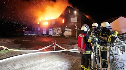 Drei Feuerwehrleute stehen vor einem Haus in Schleiden-Herhahn, das in Vollbrand steht.