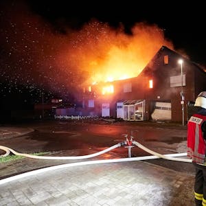 Drei Feuerwehrleute stehen vor einem Haus in Schleiden-Herhahn, das in Vollbrand steht.