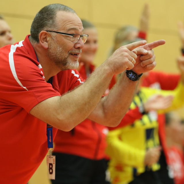 Andreas Kunzke deutet von der Trainerbank aus in Richtung Spielfeld. Im Hintergrund feiern mehrere Handballerinnen den Sieg ihrer Mannschaft.