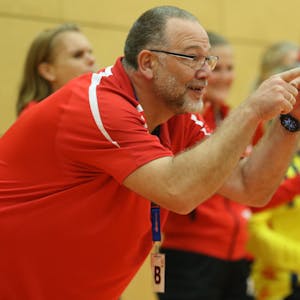Andreas Kunzke deutet von der Trainerbank aus in Richtung Spielfeld. Im Hintergrund feiern mehrere Handballerinnen den Sieg ihrer Mannschaft.