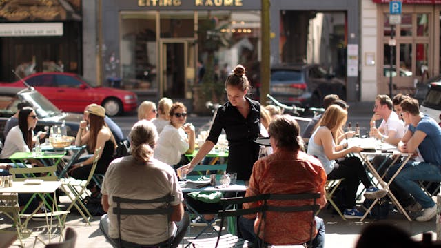 Menschen sitzen in einer Straße in der Außengastronomie an Tischen. Eine Kellnerin bedient sie.