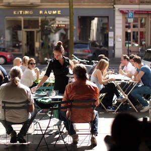 Menschen sitzen in einer Straße in der Außengastronomie an Tischen. Eine Kellnerin bedient sie.