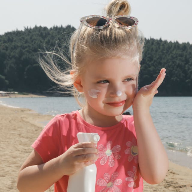 Kleines Mädchen steht an einem See und cremt sich mit Sonnenmilch ein.