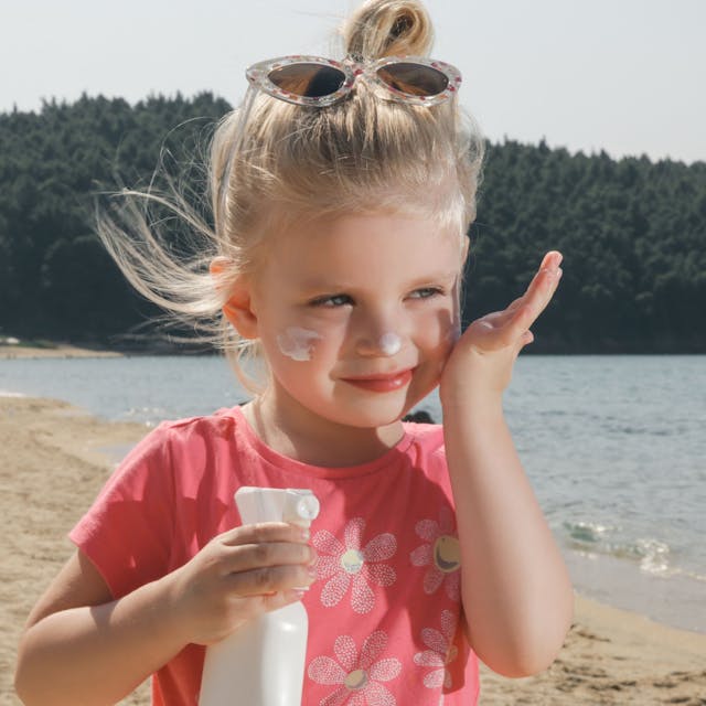 Kleines Mädchen steht an einem See und cremt sich mit Sonnenmilch ein.