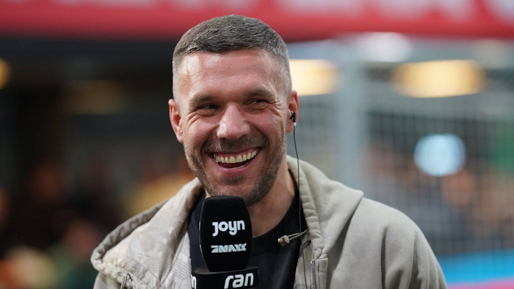 Lukas Podolski lacht mit einem Mikrofon in der Hand.&nbsp;