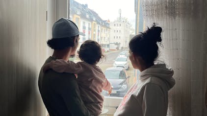 Silhouette einer dreiköpfigen Familie: Vater mit Tochter auf dem Arm, Mutter rechts, gucken aus dem Fenster