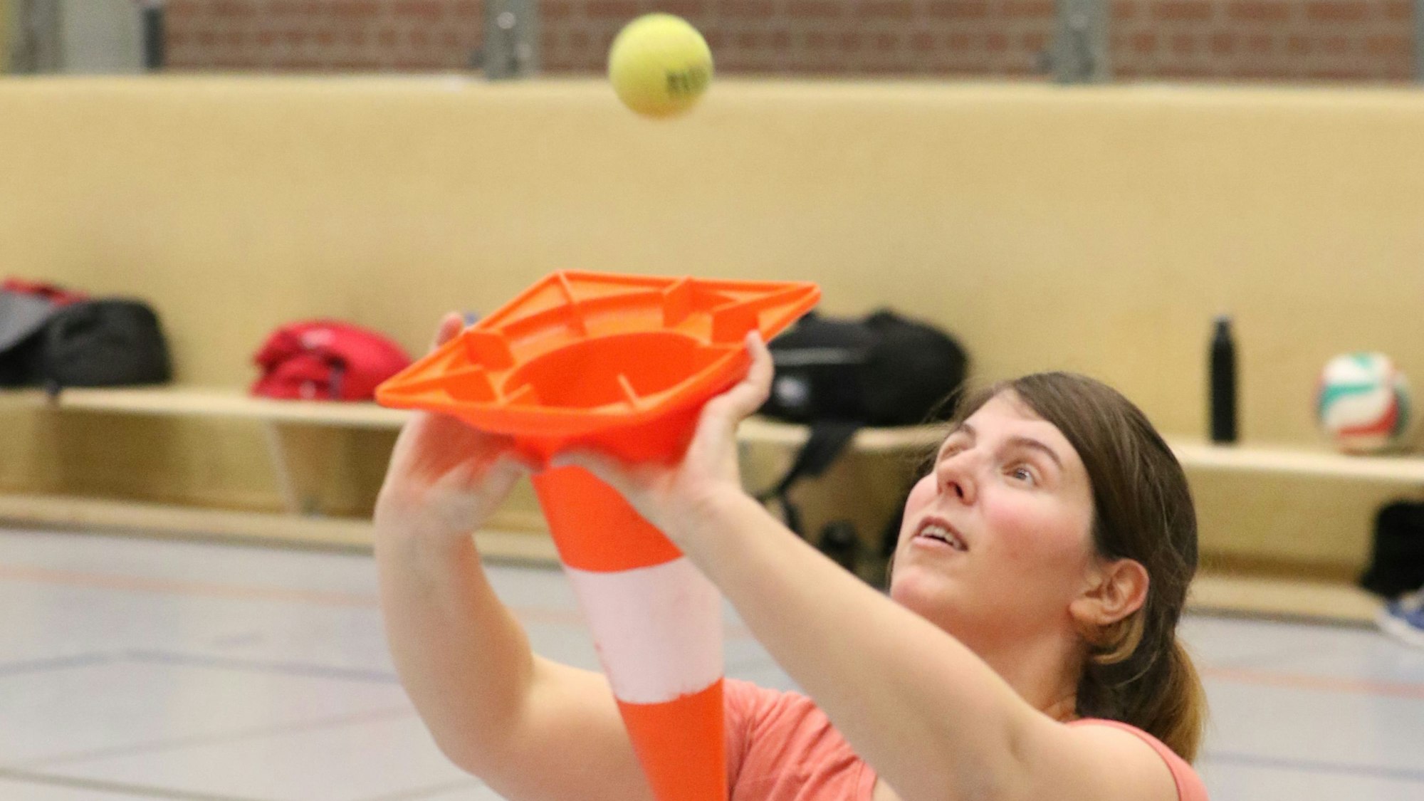 Eine Volleyballerin trainiert in einer Sporthalle das Pritschen. Sie muss mit einem Hütchen einen Tennisball fangen.