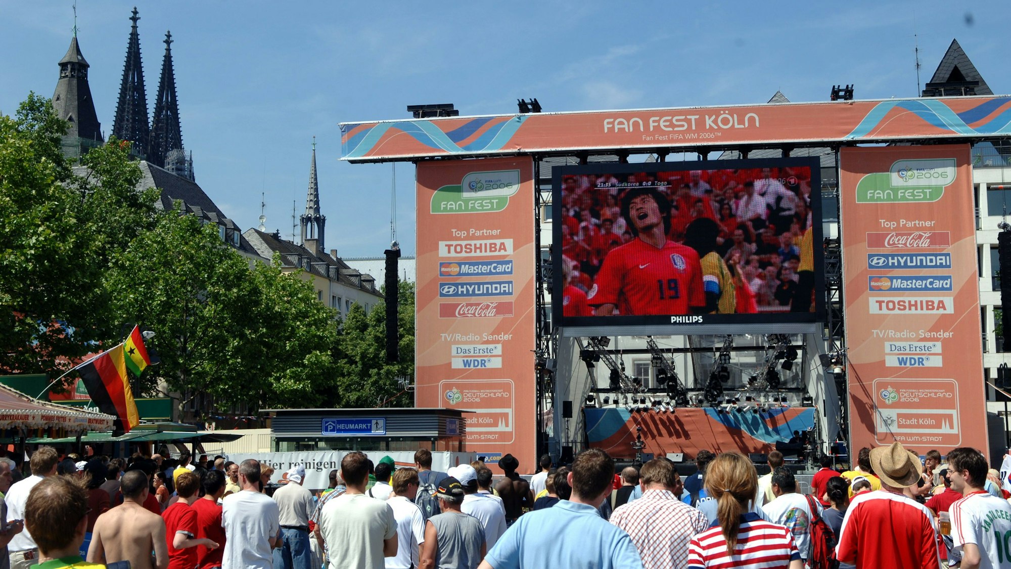 Eine große Bühne ist auf dem Heumarkt aufgebaut. Auf einer Videoleinwand wird ein Fan bei einem Fußballspiel gezeigt, darüber steht „Fan Fest Köln“.