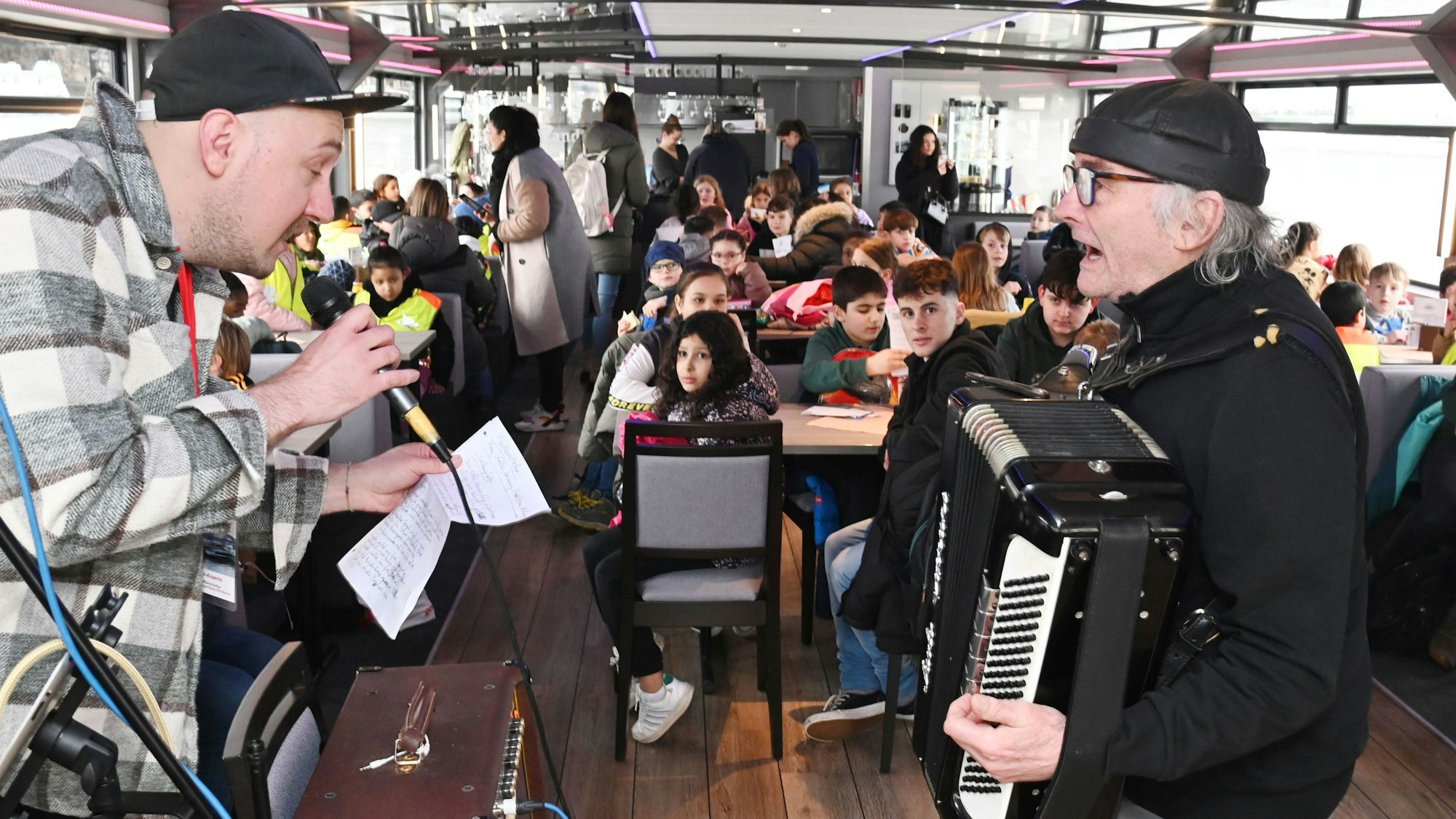 Ein Mann mit Mikrofon in der Hand und ein Mann mit Akkordeon stehen sich gegenüber. Im Hintergrund sind Kinder zu sehen, die an Tischen sitzen.