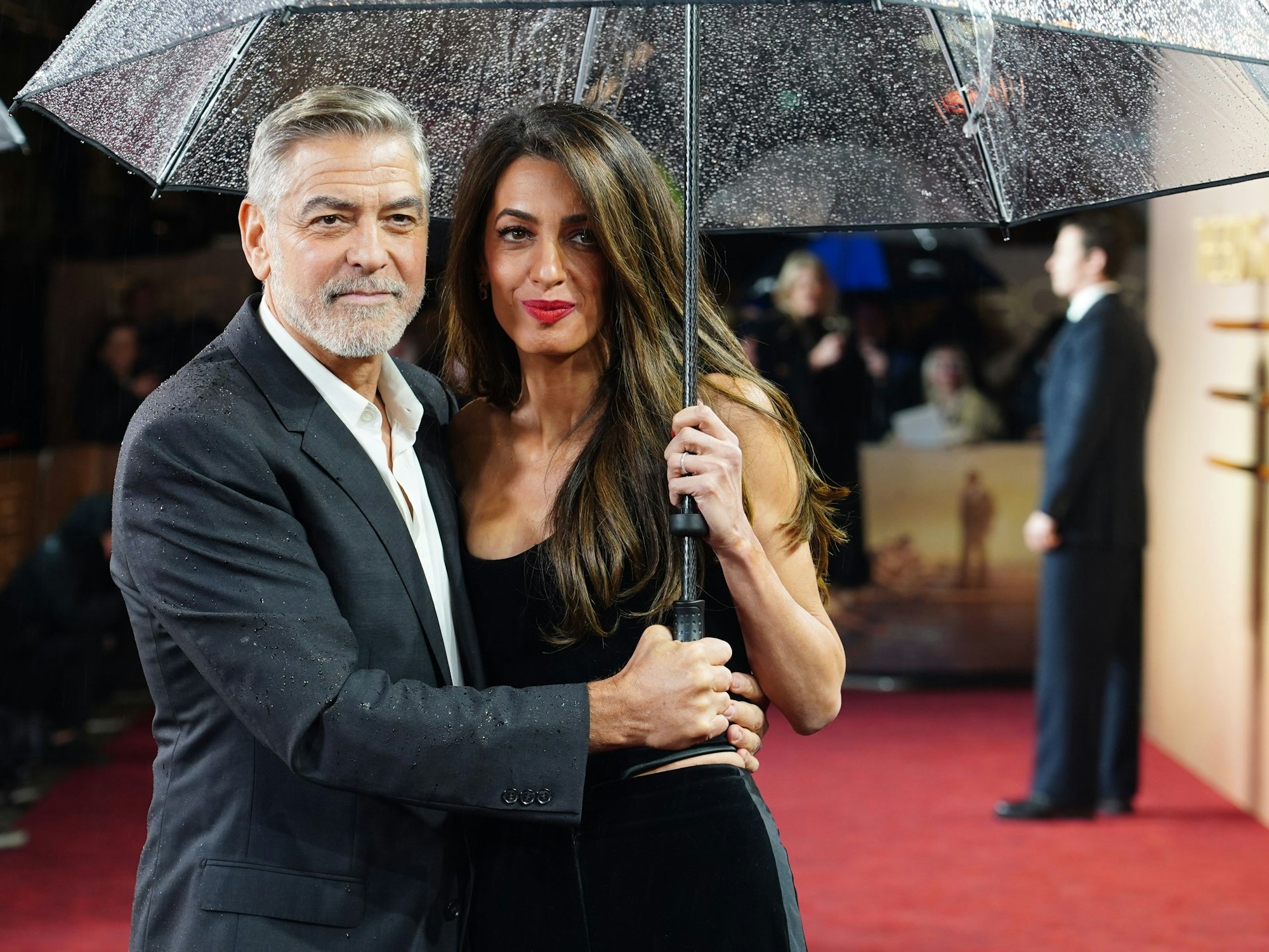George Clooney, Schauspieler aus den USA, kommt mit Ehefrau Amal Clooney unterm Regenschirm zur britischen Sondervorführung von «The Boys in the Boat» im Curzon Mayfair.