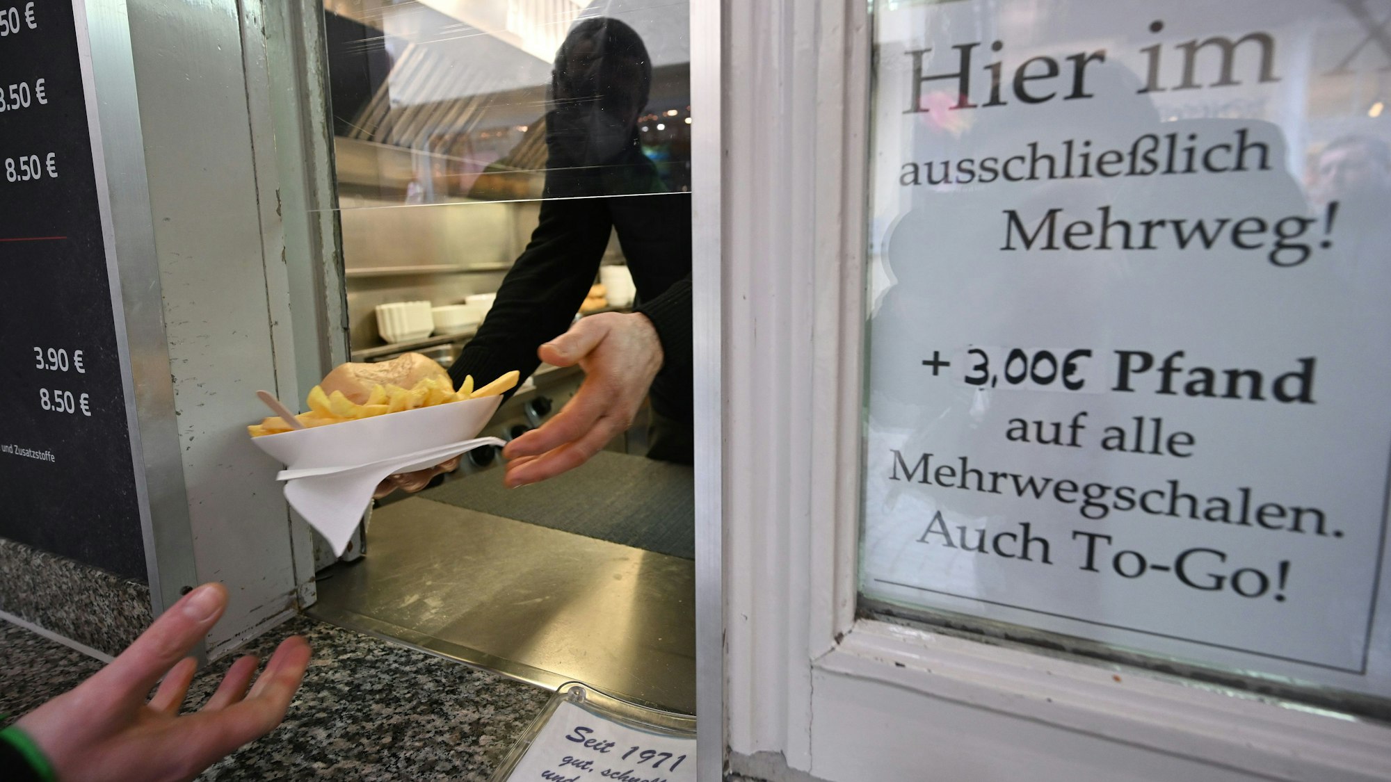 Eine Hand hält an einem Imbiss einen Mehrwegteller mit Pommes und einem Burger.