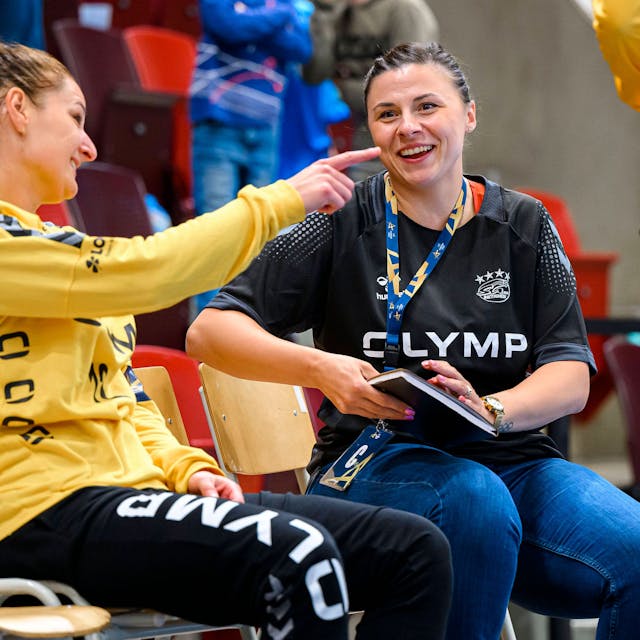 Jasmina Rebmann-Jankovic sitzt lachend neben der Bietigheimer Torhüterin Melinda Szikora, die ihr was erzählt.