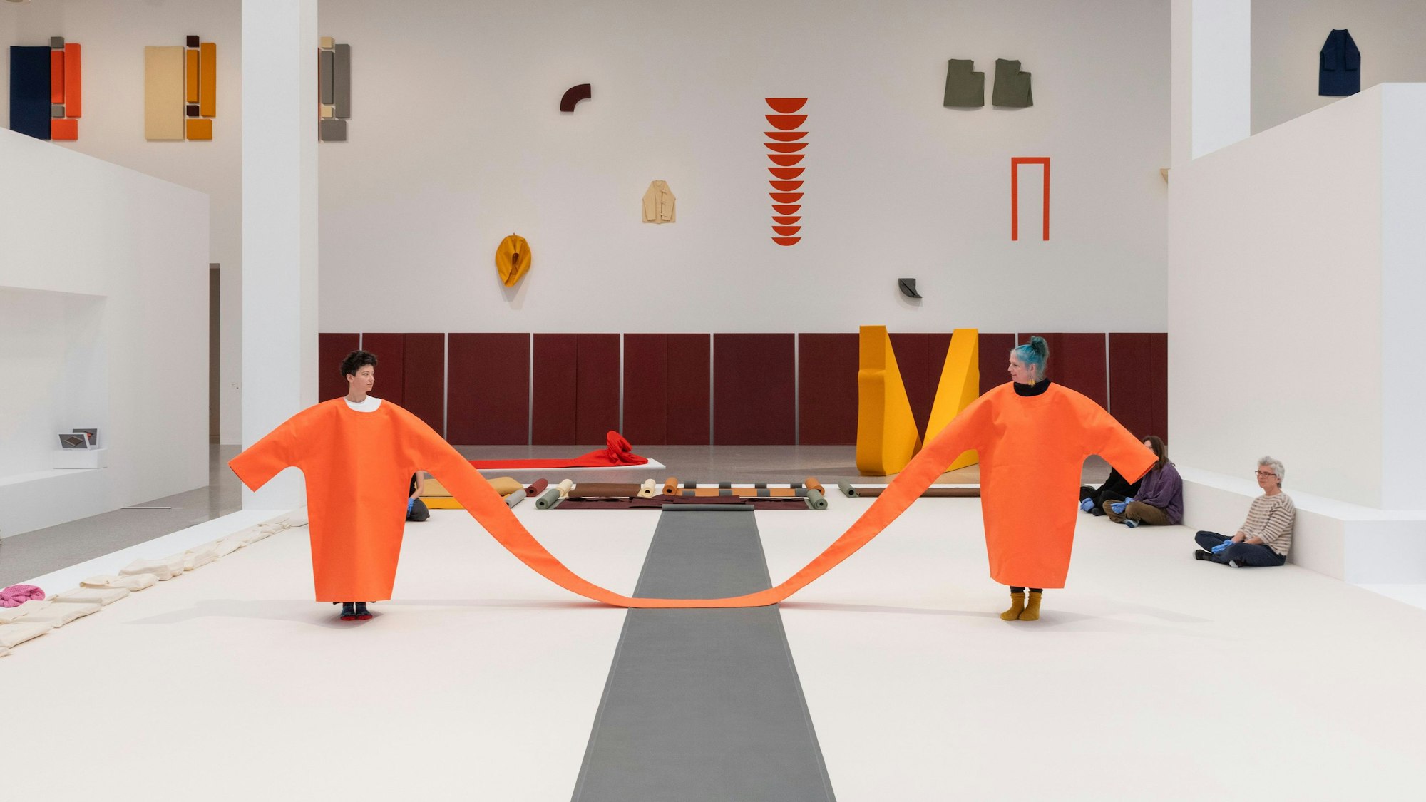 Zwei Menschen stehen in orangefarbenen Kleidern im Raum. Die Kleider sind an einem Ärmel zusammengenäht.