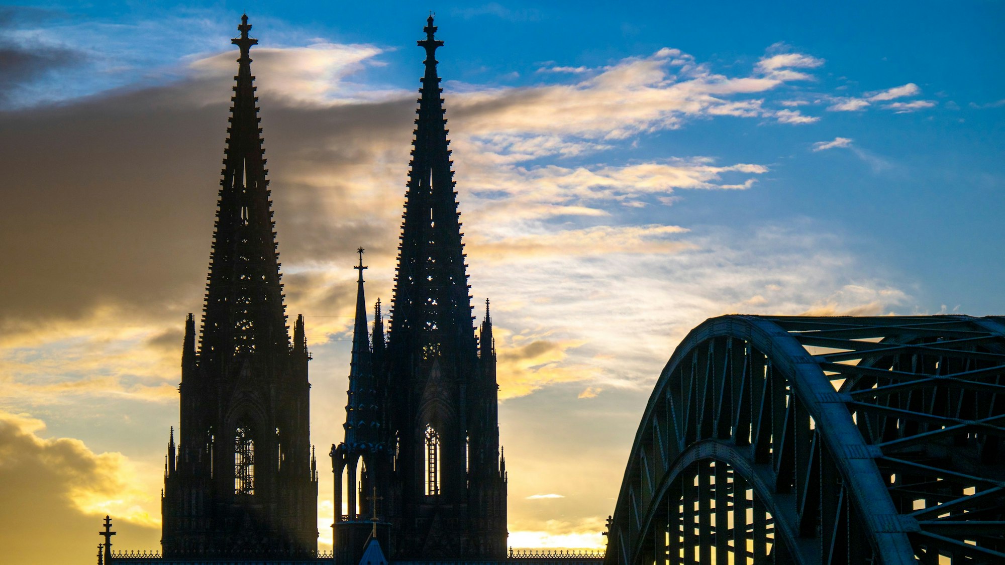 Die Sonne geht hinter dem Kölner Dom und der Hohenzollernbrücke unter, mehrere Wolken schieben sich vor den blauen Himmel. (Symbolbild)