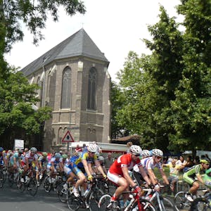 Rennradfahrer fahren im Pulk, im Hintergrund ist eine Kirche zu sehen.