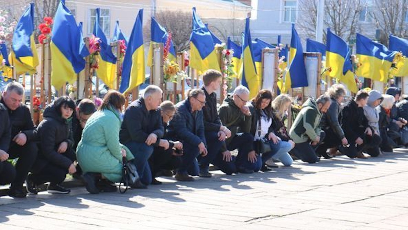 Das Foto zeigt knieende Menschen und dahinter viele ukrainische Flaggen.