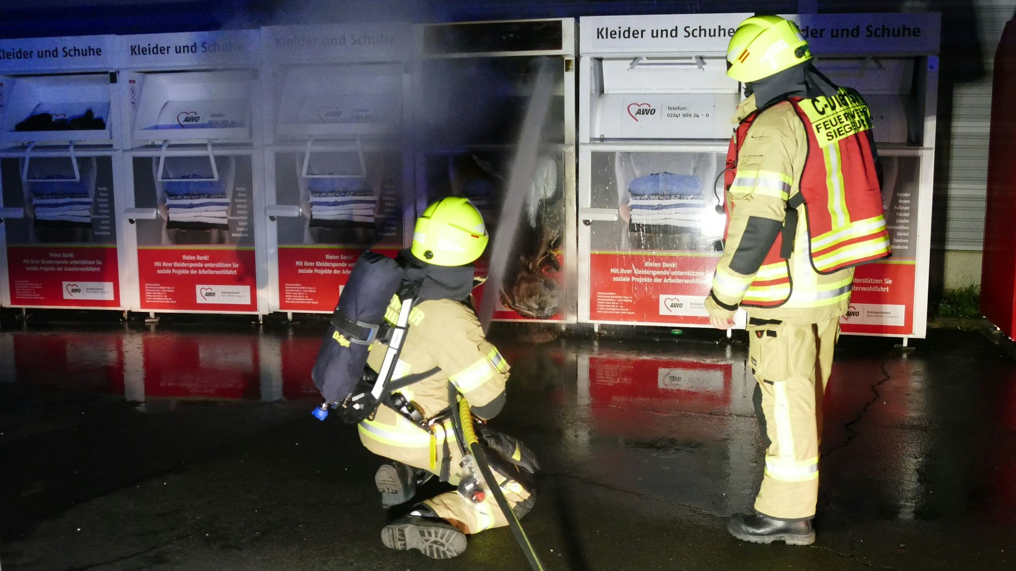 Zwei Feuerwehrleute vor einem brennenden Altkleidercontainer. Eine Person kniet und richtet den Wasserstrahl auf den Sammelbehälter.