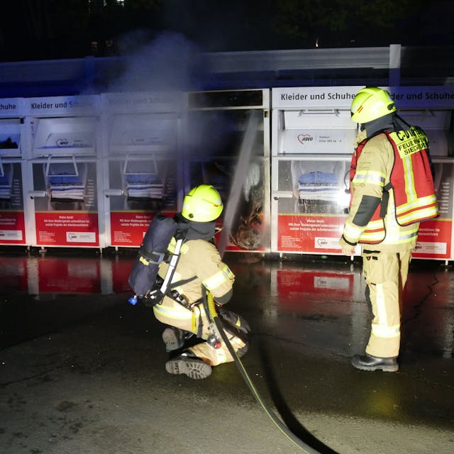 Zwei Feuerwehrleute vor einem brennenden Altkleidercontainer. Eine Person kniet und richtet den Wasserstrahl auf den Sammelbehälter.