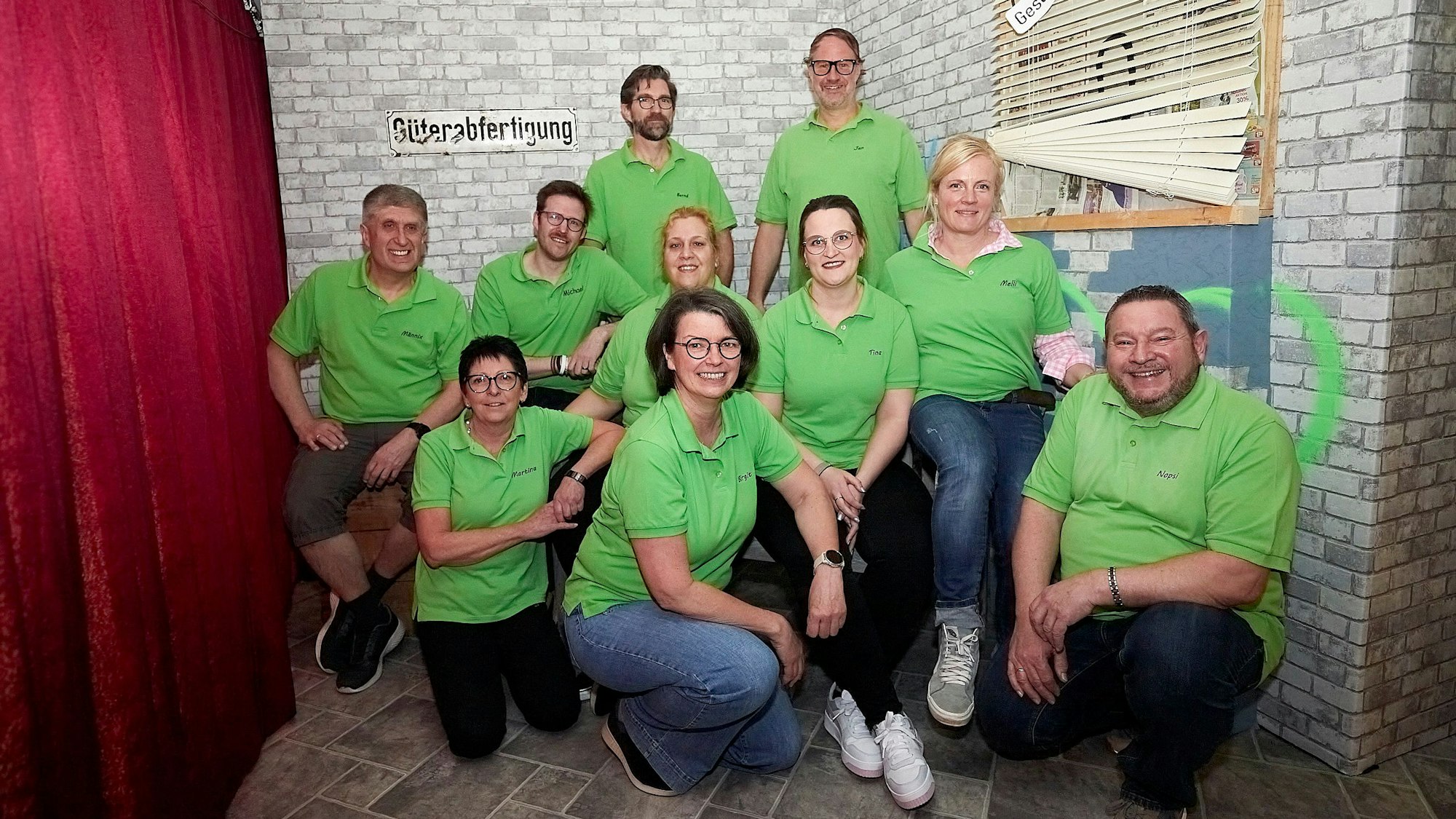 Einige Frauen und Männer sind in grünen T-Shirts in einer Theaterkulisse zu sehen. Sie gehören zur Kleinen Theaterschar Kronenburg und bereiten sich auf ihre Aufführung vor.