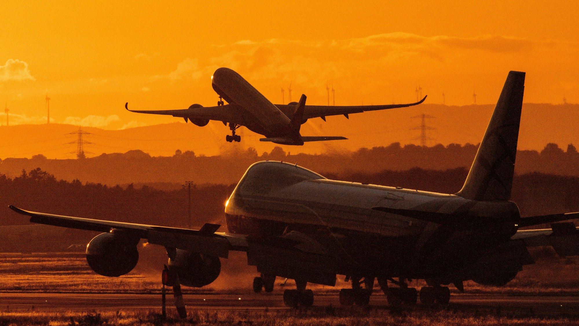 Ein Flugzeug steht bei Sonnenuntergang auf einem Rollfeld. Im Hintergrund hebt eine anderes Flugzeug ab