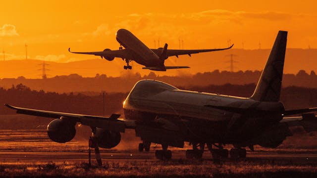 Ein Flugzeug steht bei Sonnenuntergang auf einem Rollfeld. Im Hintergrund hebt eine anderes Flugzeug ab