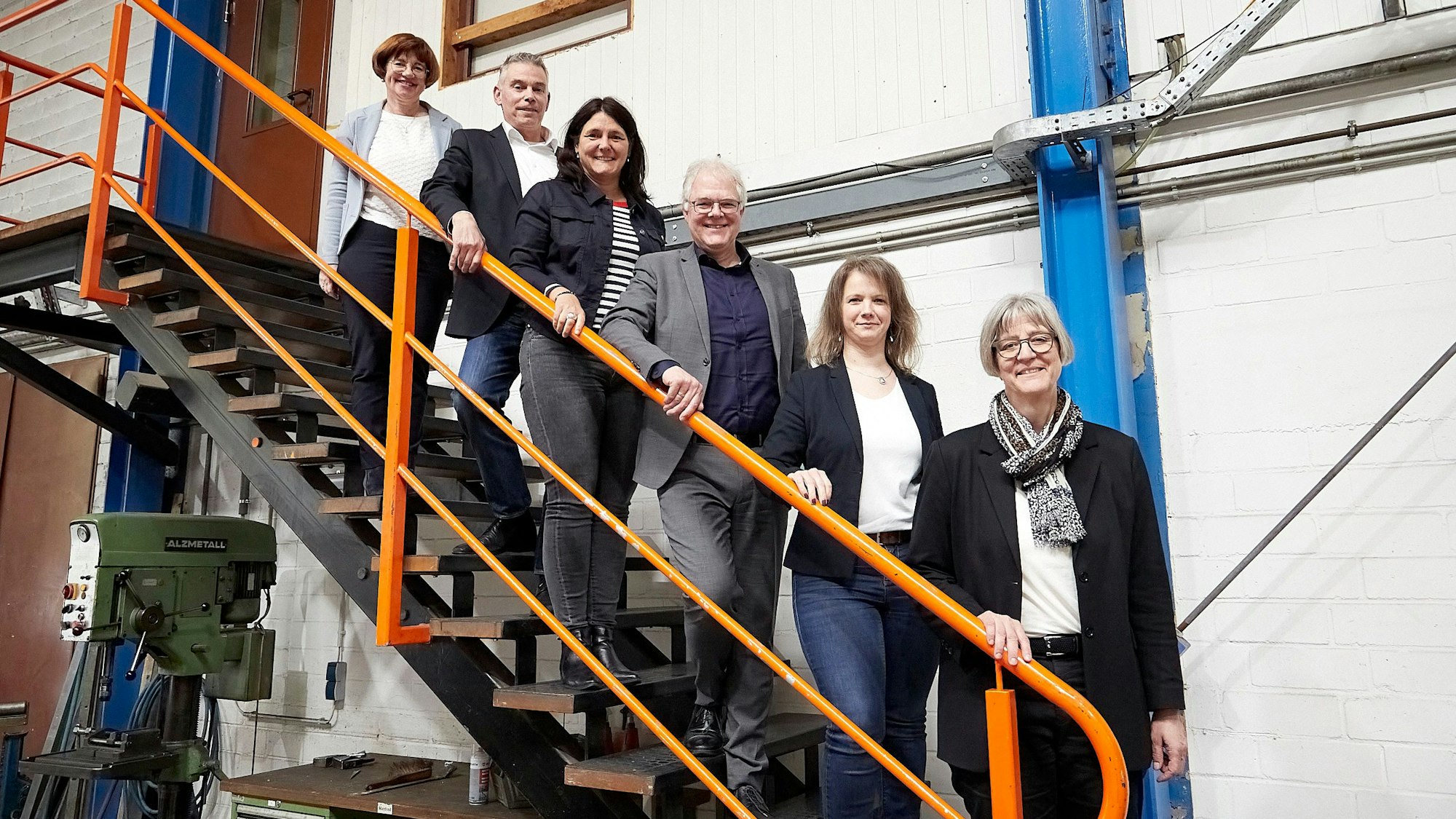 Vier Frauen und sechs Männer stehen in einer Produktionshalle auf einer Treppe. Die meisten legen eine Hand auf das orangefarbene Geländer.