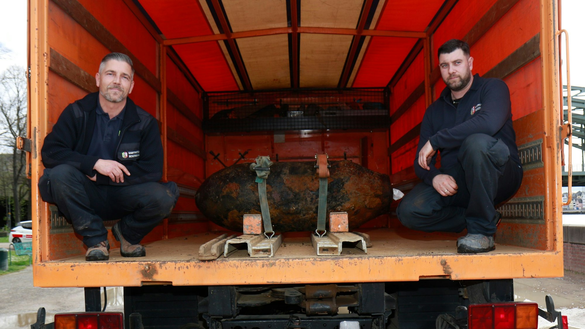 Zwei Männer in Dienstkleidung im Innenraum eines Lastwagens, in ihrer Mitte ist eine entschärfte Bombe festgeschnallt