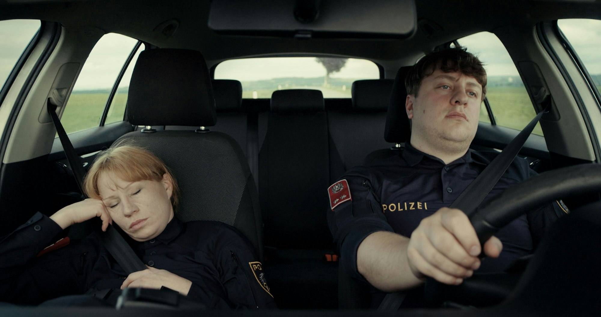 Zwei Polizisten fahren im Streifenwagen. Die Frau schläft, der Mann am Steuer langweilt sich.