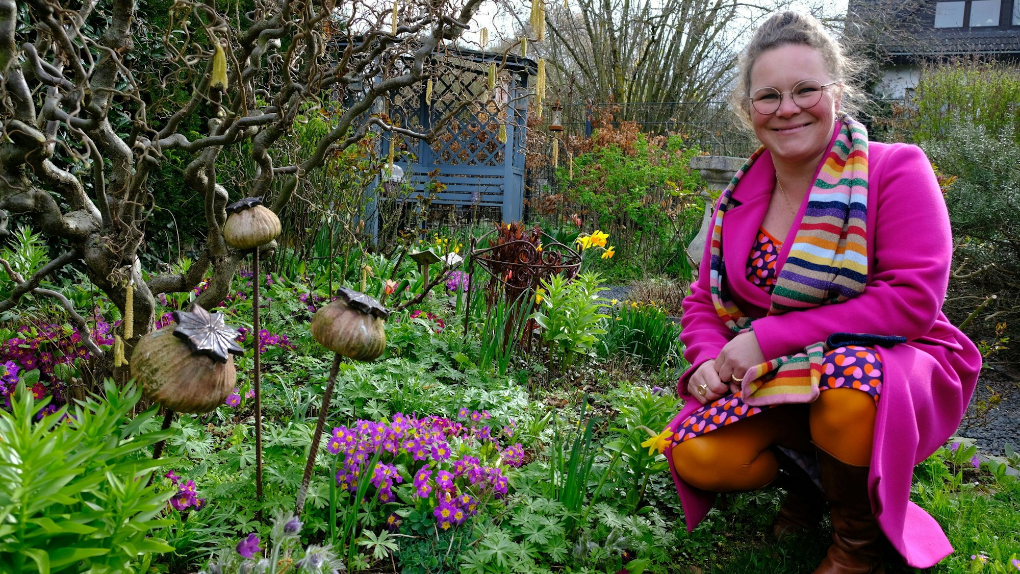 Kai Judith Wetzel kniet an einem Blumenbeet in ihrem Garten. Sie trägt einen pinkfarbenen Mantel, ein buntes Kleid und einen gestreiften Schal.