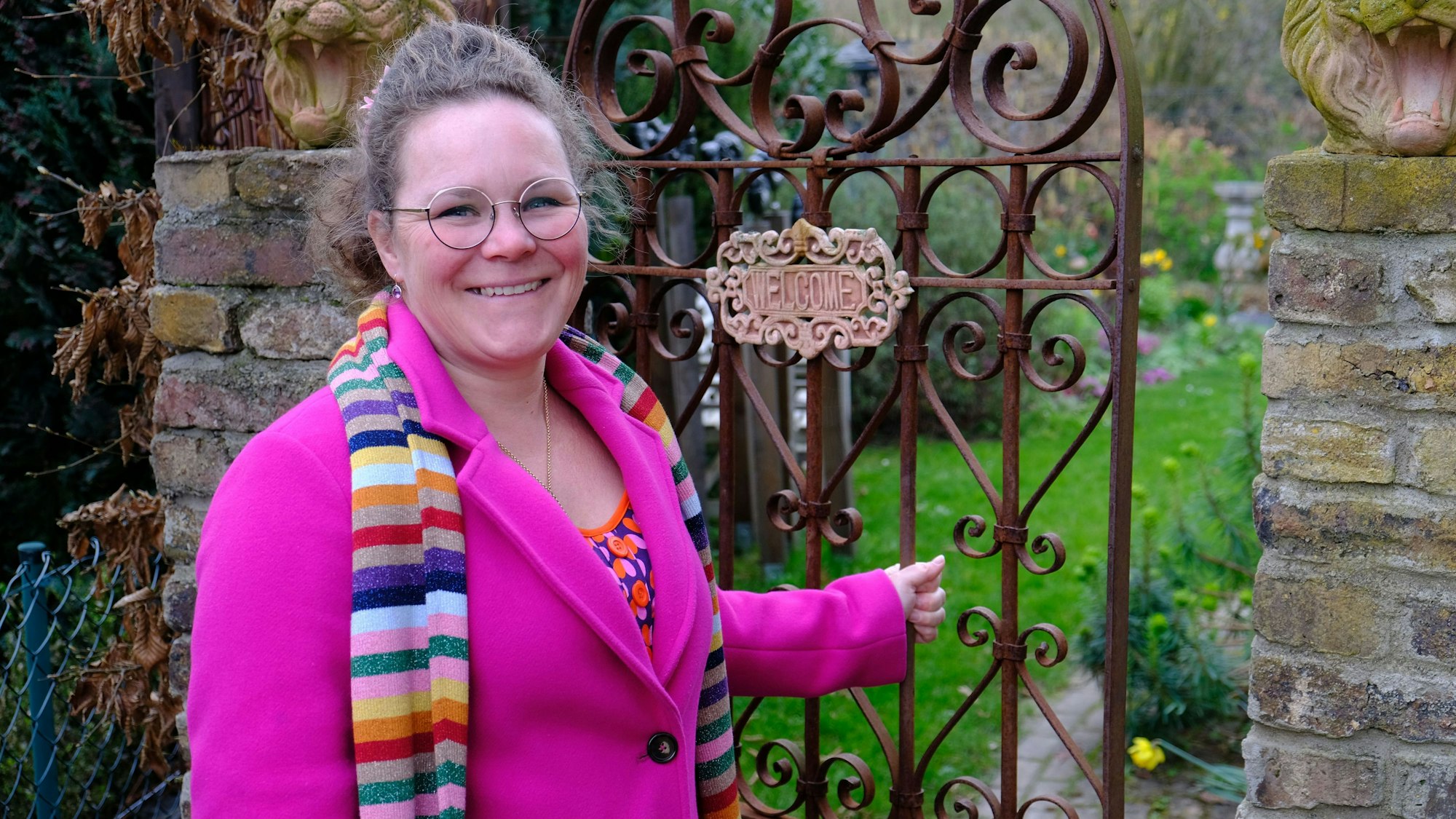 Kai Judith Wetzel steht in einem pinkfarbenen Mantel und mit einem bunt gestreiften Schal an der schmiedeeisernen Pforte ihres Gartens.