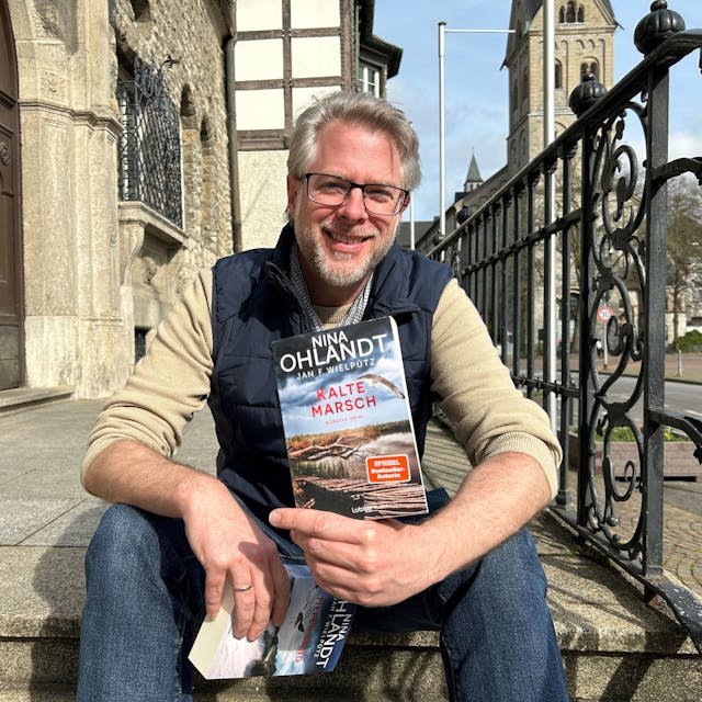 Autor Jan Wielpütz sitzt auf der Rathaustreppe in Bergisch Gladbach und zeigt seinen neuen Krimi „Kalte Marsch“&nbsp;