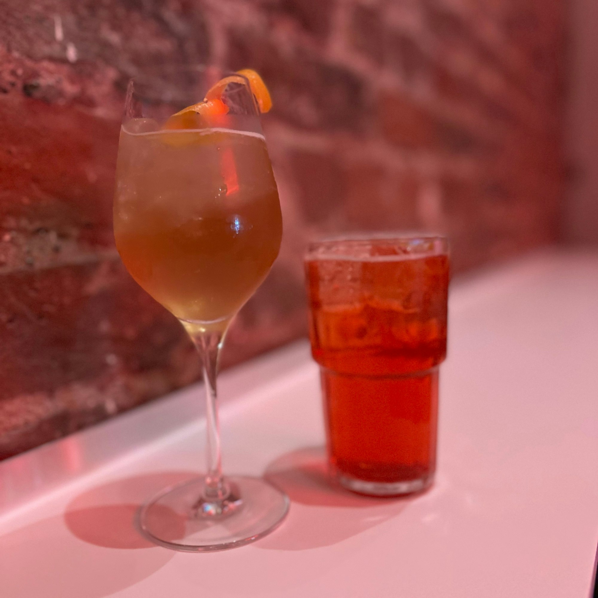 Ein Weiglas mit einem Longdrink in Orange und ein Glas mit einem roten Longdrink vor einr Ziegelwand