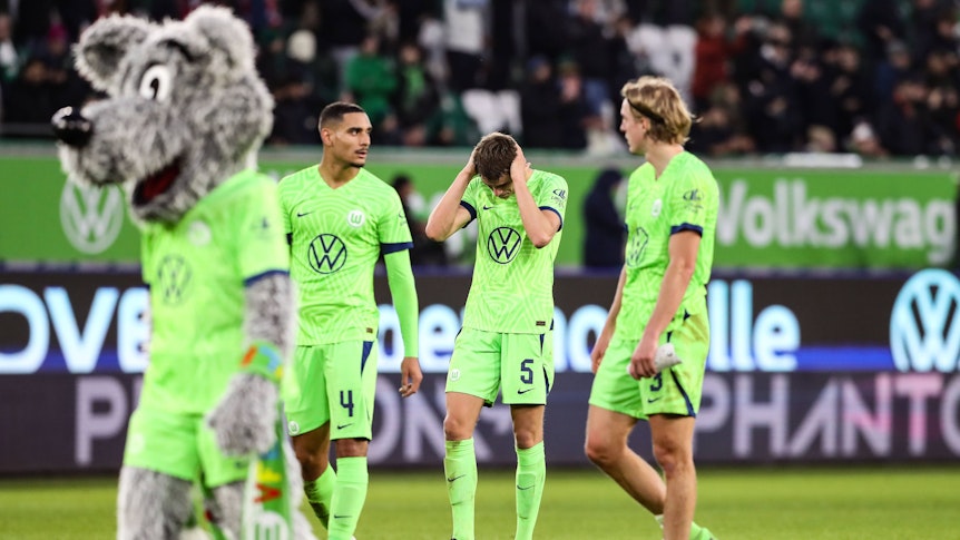 Drei Wolfsburg-Spieler ärgern sich. Auf der linken Seite läuft das Maskottchen Wölfi über den Platz.