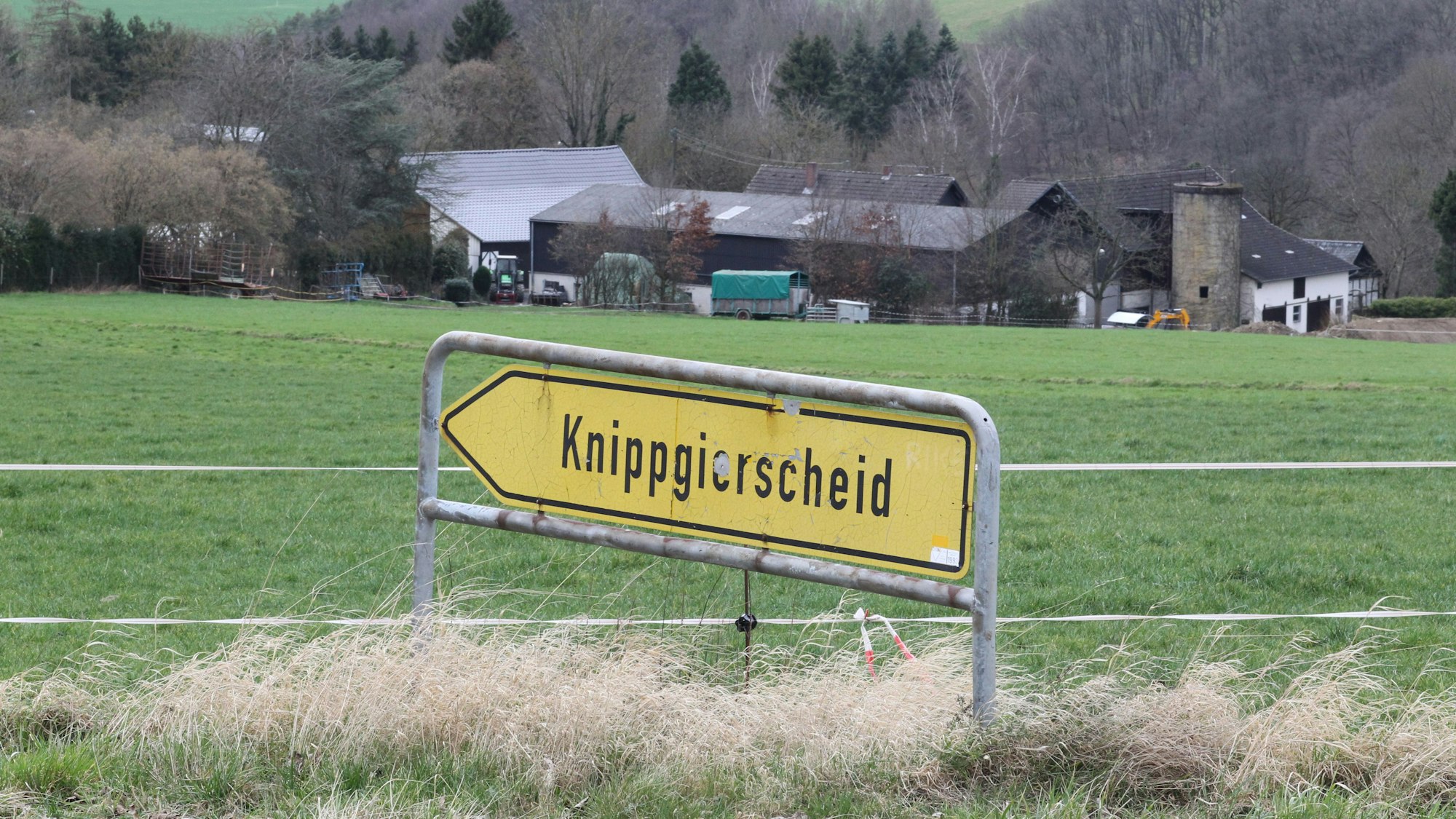 Blick über ein landwirtschaftliche Wiese auf ein Gehöft mit einem Schild im Vordergrund, das den Weg zum Dorf Knippgierscheid zeigt.
