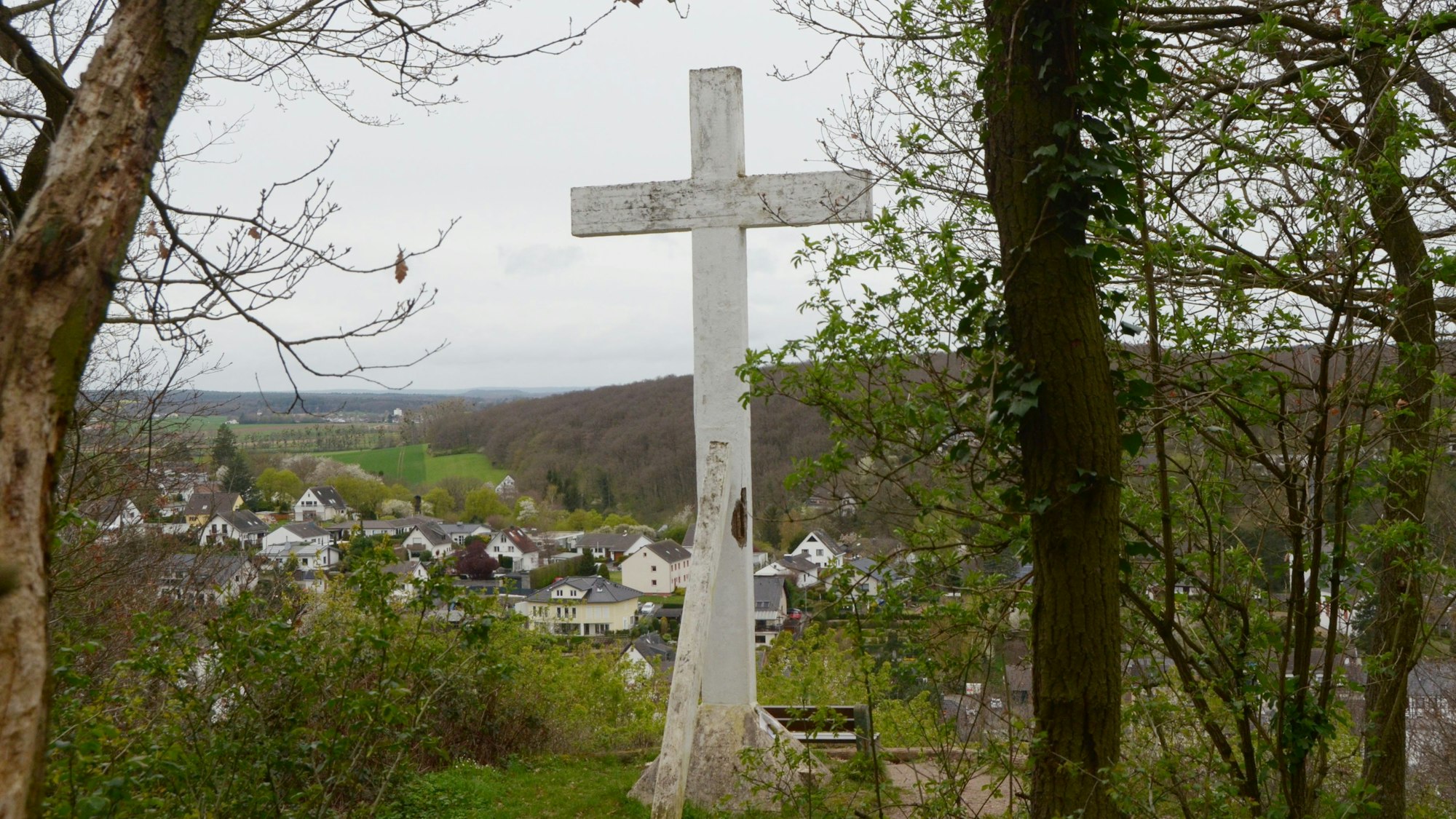 Das Kreuz steht am Waldrand, davor ist eine Sitzbank platziert. Im Bildhintergrund das Erfttal und der Ort Kreuzweingarten.