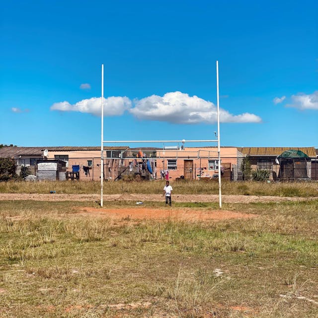 Ein Kind steht vor den Stangen eines Rugby-Tors auf einem Sportplatz in Südafrika. Im Hintergrund sind die Häuser einer Township zu sehen.