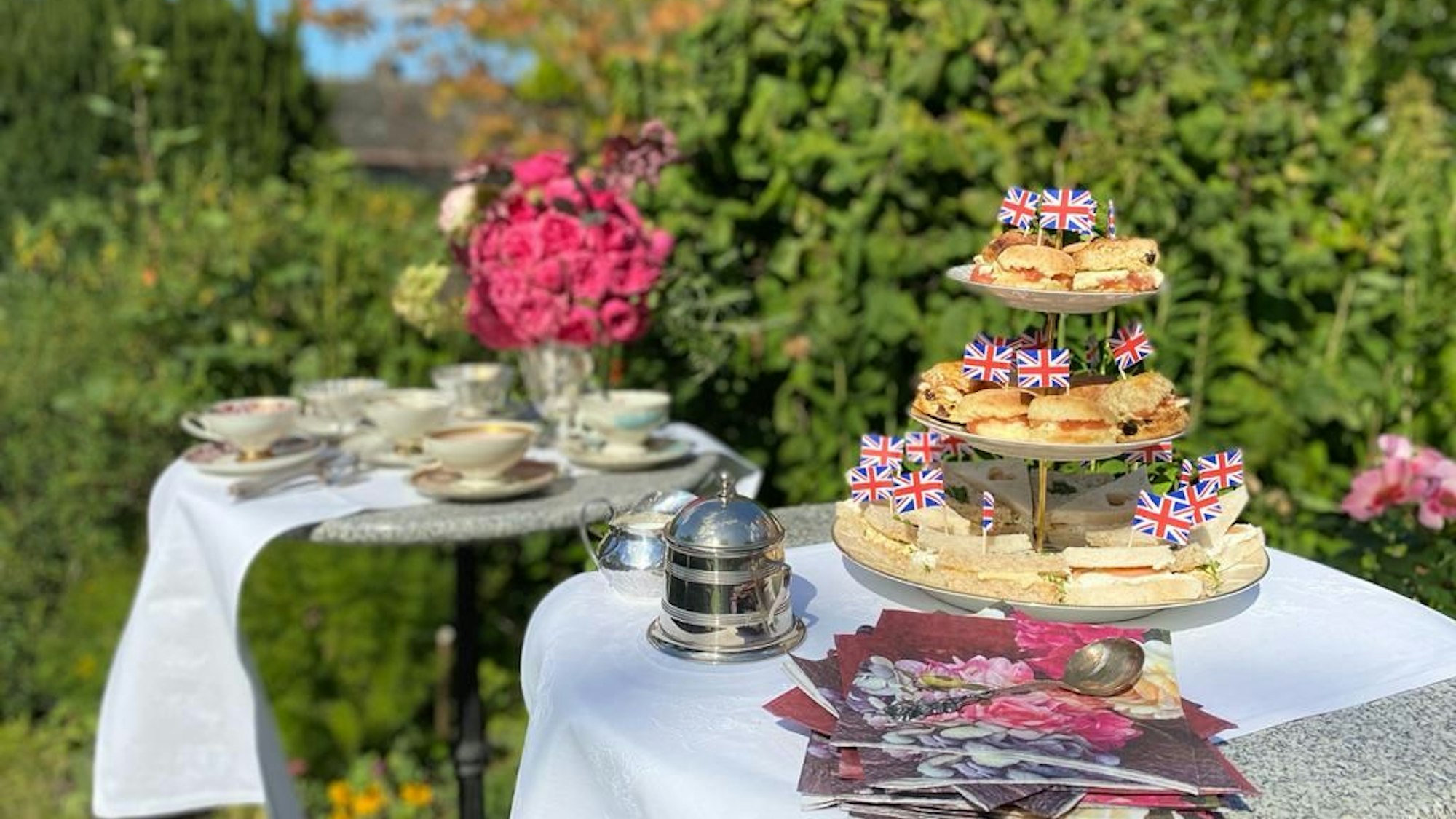Auf zwei Tischen im Garten stehen auf weißen Tischdecken Teetassen, Blumen sowie Sandwiches und Scones, dekoriert mit der britischen Flagge.