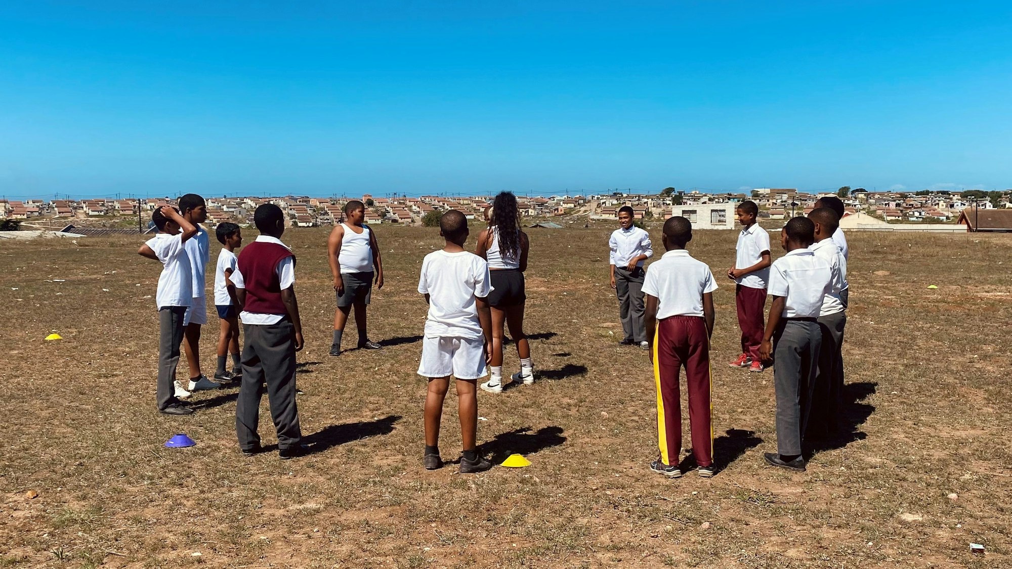 Eine Gruppe von Schülern steht auf dem Gelände einer südafrikanischen Schule im Kreis und umringt eine Freiwillige, die Rugby-Übungen anleitet.