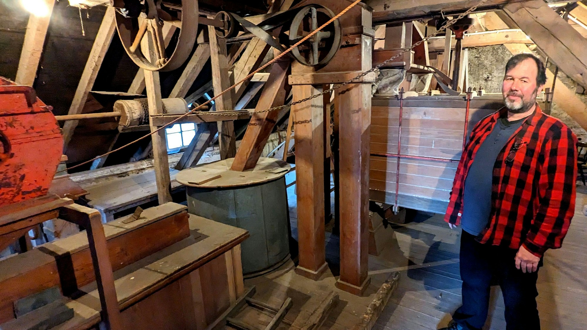 Wolfgang Pütz zeigt die alten Gerätschaften im Dachboden der Hauserbachmühle. Er trägt ein rot-kariertes Holzfällerhemd.