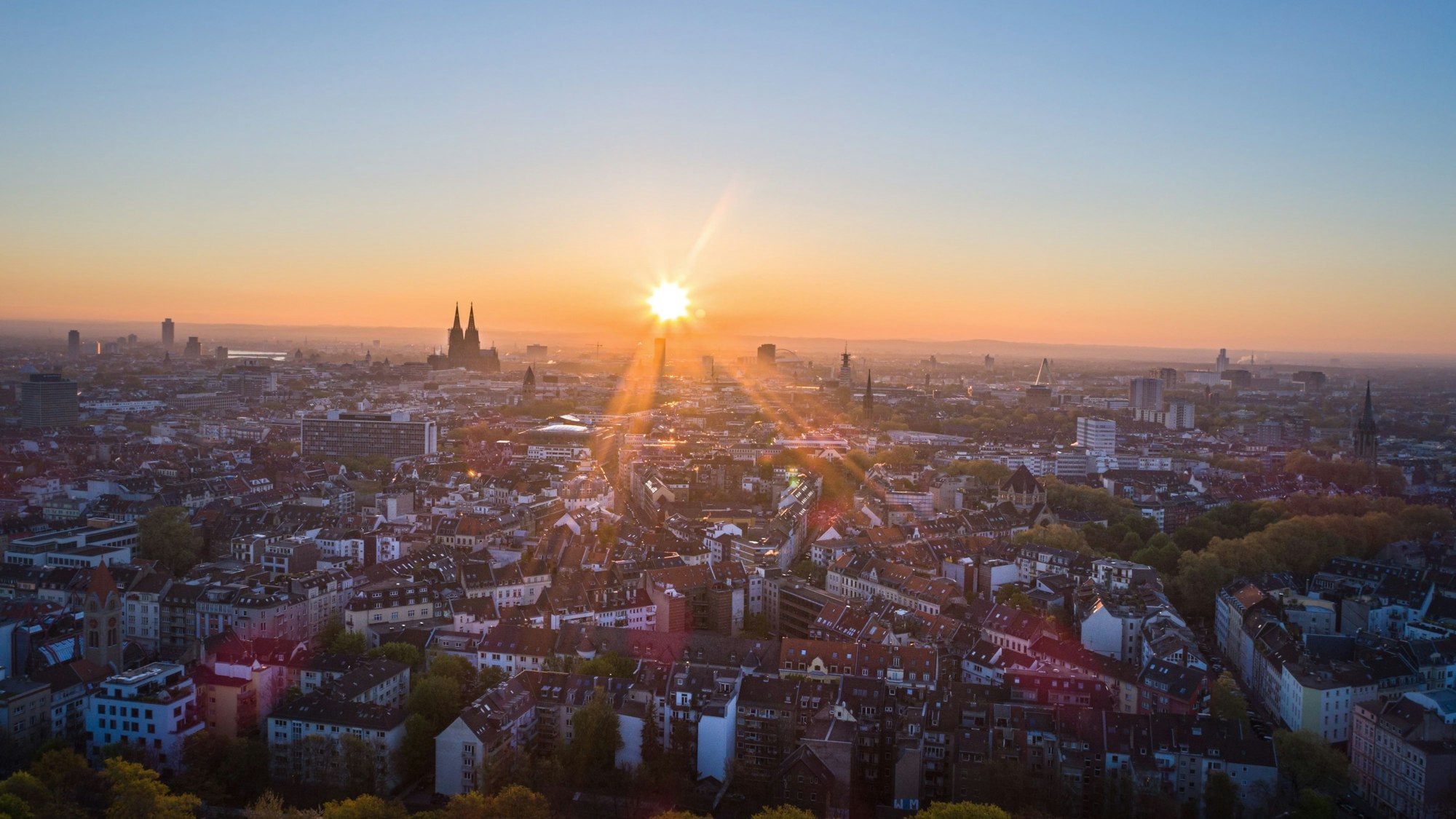Die Sonne geht an einem wolkenlosen Morgen mit blauem Himmel neben dem Kölner Dom (Hohe Domkirche St. Petrus) über dem Stadtzentrum auf.