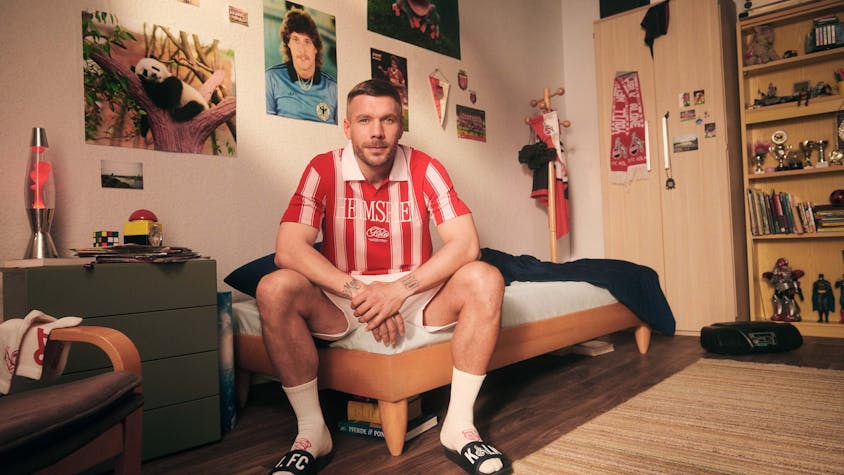 Lukas Podolski sitzt in einem Jugendzimmer auf dem Bett. Er trägt ein rot-weiß gestreiftes Poloshirt.