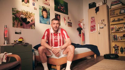Lukas Podolski sitzt in einem Jugendzimmer auf dem Bett. Er trägt ein rot-weiß gestreiftes Poloshirt.