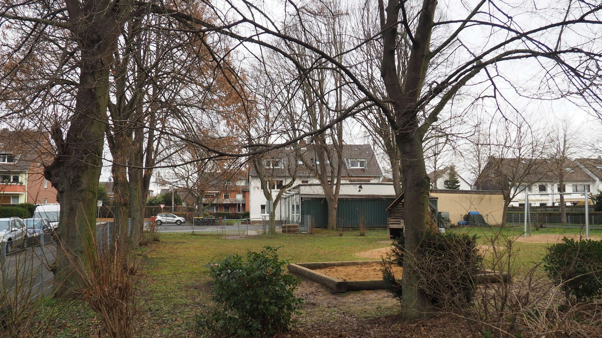 Im Hintergrund das Feuerwehrgerätehaus, im Vordergrund der kleine Park mit alten Bäumen und einem Sandkasten.