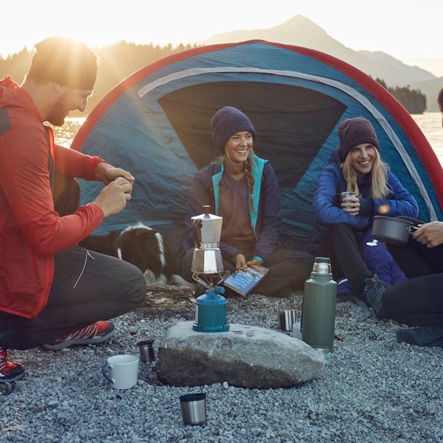 Eine Trekking-Gruppe kocht vor einem Zelt Kaffee auf einem Gaskocher.