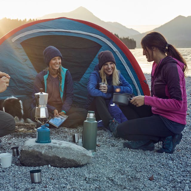 Eine Trekking-Gruppe kocht vor einem Zelt Kaffee auf einem Gaskocher.