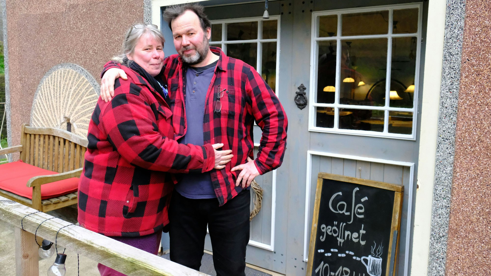 Karin Goebel und Wolfgang Pütz stehen vor dem Eingang ihres Cafés. Beide tragen rot-karierte Holzfällerhemden.