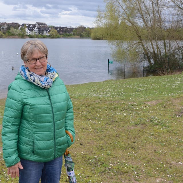 Eine Frau mit grüner Daunenjacke steht am Ufer eines Sees. Im Hintergrund sind Häuser jenseits des Sees zu sehen.