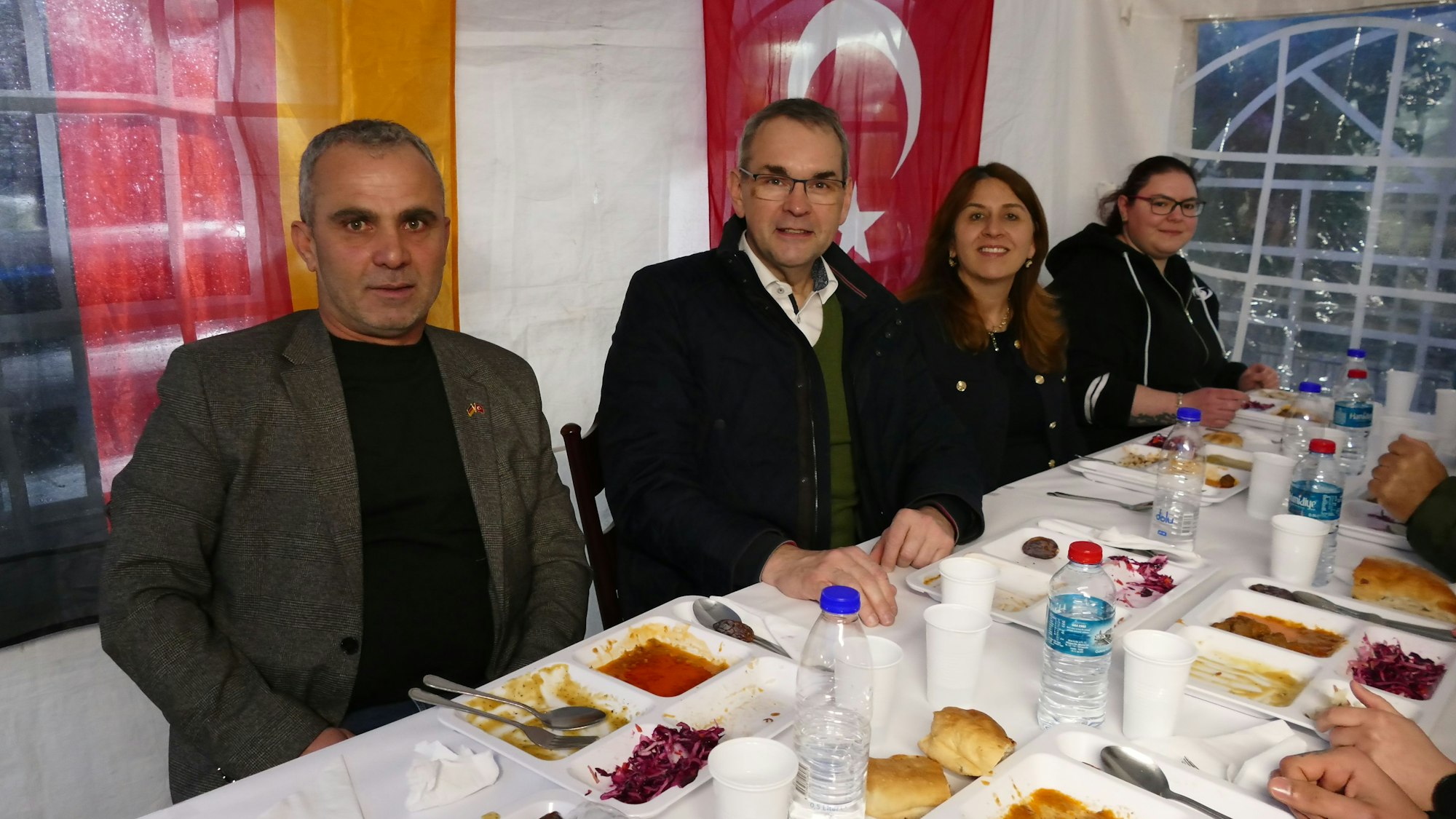 Zwei Männer und zwei Frauen sitzen vor Speisen an einem gedeckten Tisch. Hinter ihnen hängen eine türkische und eine deutsche Fahne.
