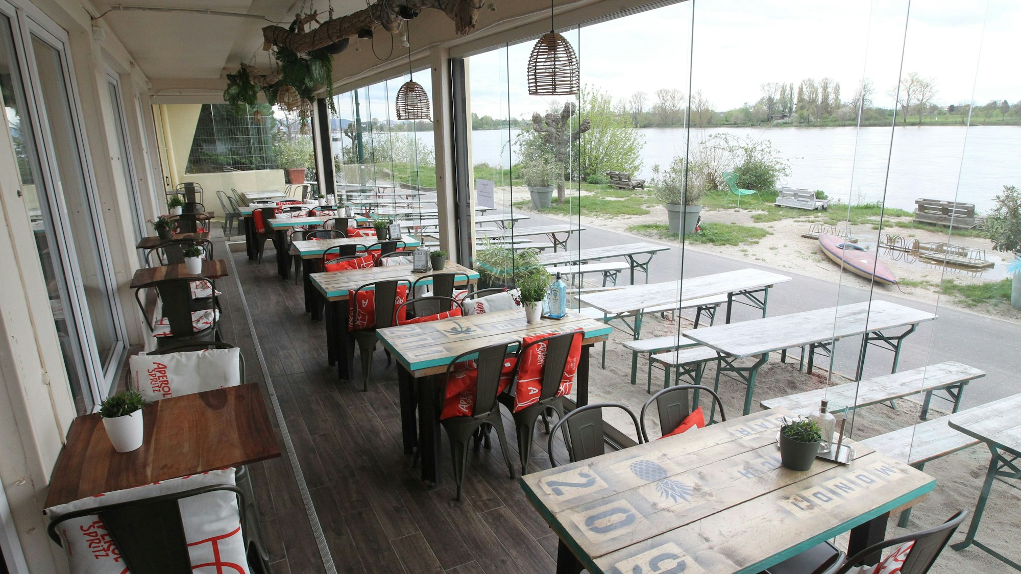 Tische stehen auf einer überdachten Terrasse, ein Sandstrand und der Rhein liegen daneben.