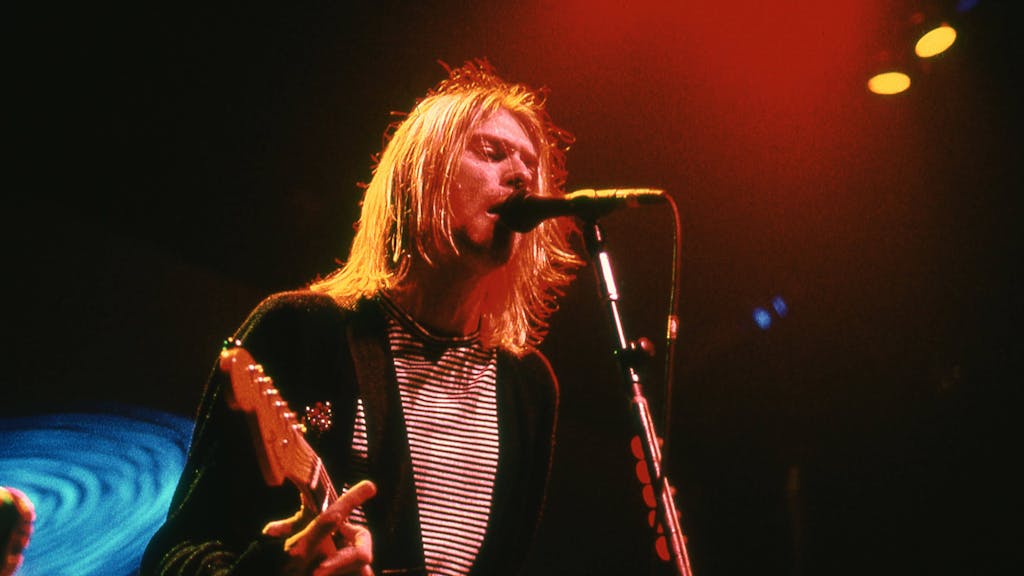 Kurt Cobain steht mit langen Haaren und seiner Gitarre vor einem Mikrofon und singt.